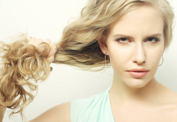 A blonde woman with curly hair, wearing a light turquoise top, looks confidently at the camera while pulling her hair to one side.