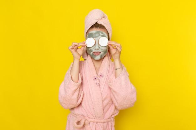 Woman applying cucumber slices over eyes while wearing face mask and bathrobe on yellow background.