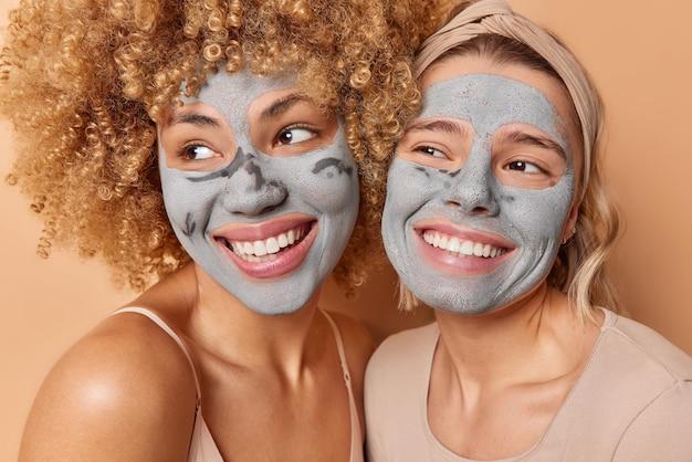 Two women smiling while wearing facial masks, standing against a soft beige background.